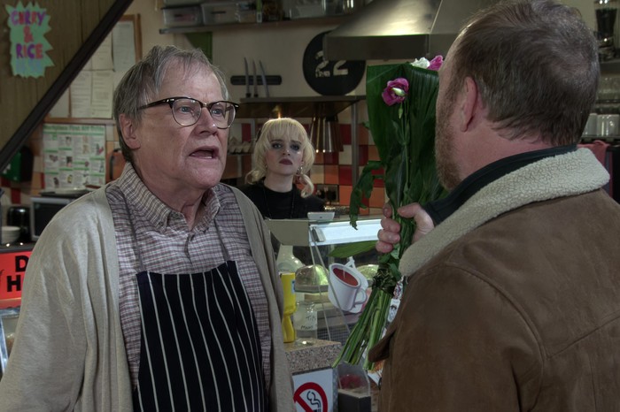 Roy chats to Mal in the cafe, with Nina peering over his shoulder from behind the counter, in a scene from Coronation Street