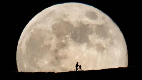 Michael Meighan Silhouette of boyish character in an anorak straddling a stationary BMX bike with an object in its front basket on a mountainside framed by the full Moon behind