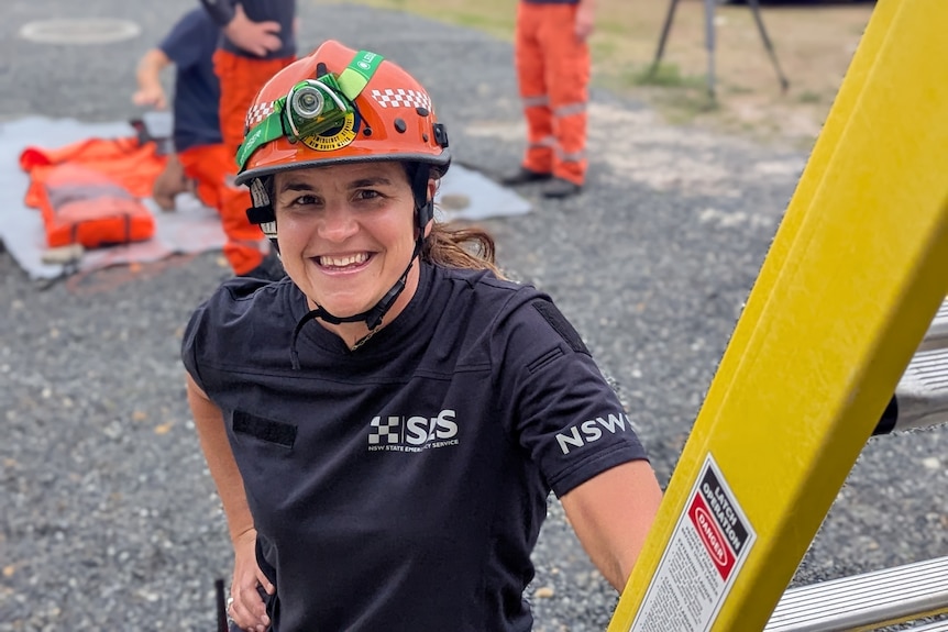 A smiling woman in a helmet leans on a ladder.