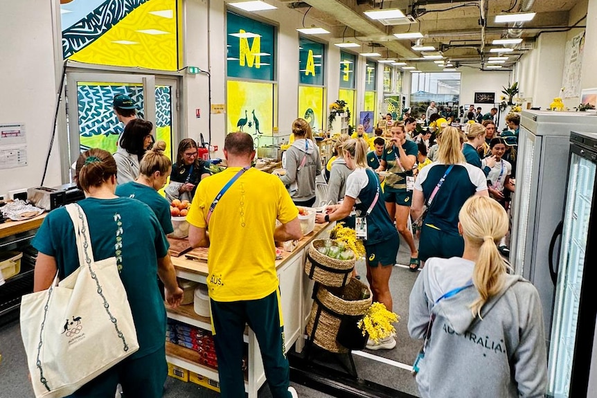 A crowd of athletes dressed in green and gold in the lobby of a building with food on offer.