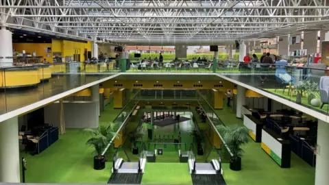 Matt Marvel/BBC The interior of a large, open-plan office building. Three floors are visible. The lower one has a modern entrance with pass barriers. Two escalators take people to the next floor, which has green flooring. The third level is a mezzanine one with yellow, green flooring, walls and desks.  The ceiling has metal struts and glass above.