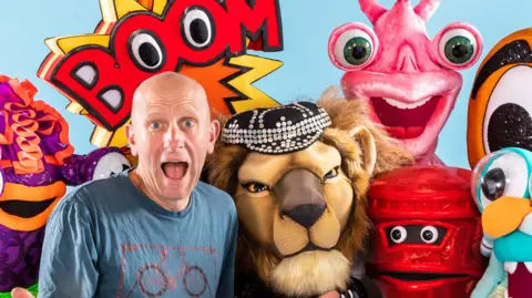 Tim Simpson Tim Simpson with a surprised face next to a variety of masks which have been used in The Masked Singer