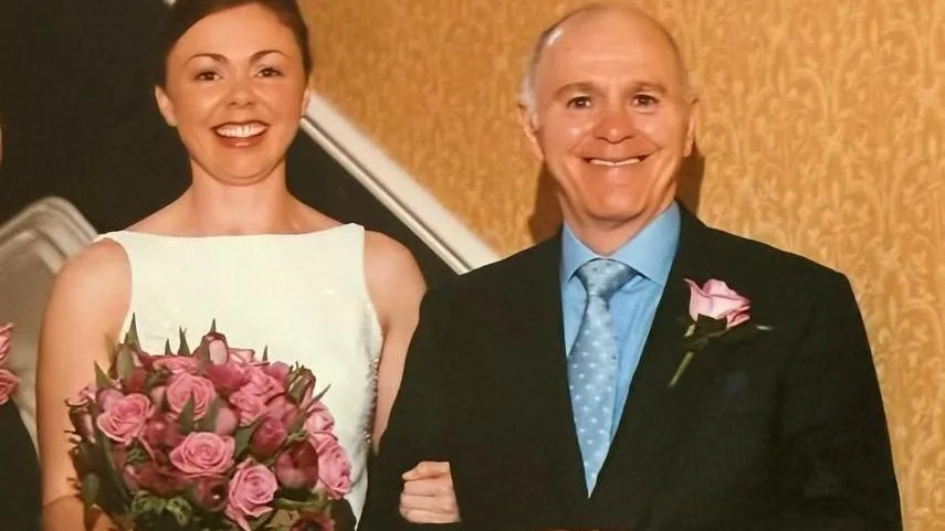 A beaming bride in a white wedding dress holding a bunch of roses and linking arms with a smiling gentleman in a smart dark suit.