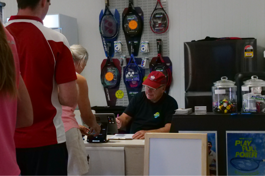 people lining up to see paul at the desk to sign in and pay, tennis racquets on the wall behind him