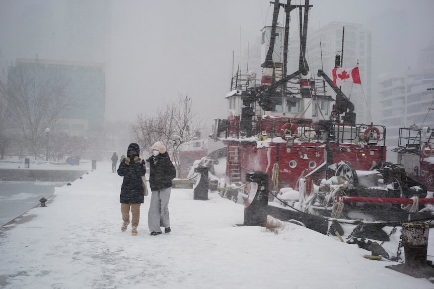 People walk next to a snow-covered boat, flying the Canadian flag, and lake.