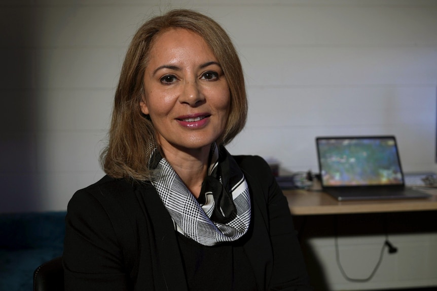 A woman with blonde hair sits and smiles, her desk and computer behind her.