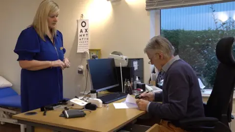 Image shows Dr Cooper writing at his desk, in a consultation room, alongside practice manager Tracey Jean. The doctor wears a blue jumper, with grey hair and glasses.