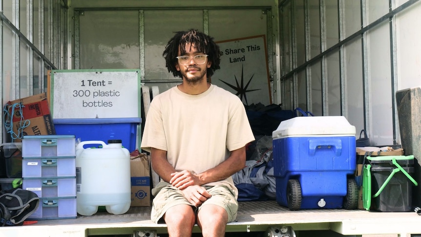 a man sitting on the back of large truck, he is wearing glasses, there is oiles of tents and camping gear behind him