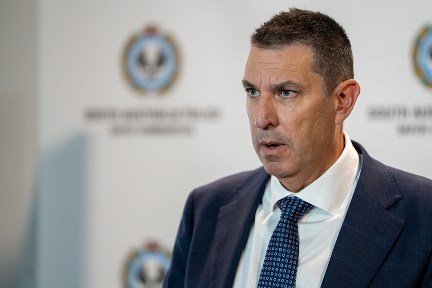 A man with short brown hair wearing a suit and blue tie looks off camera with a concerned expression. 