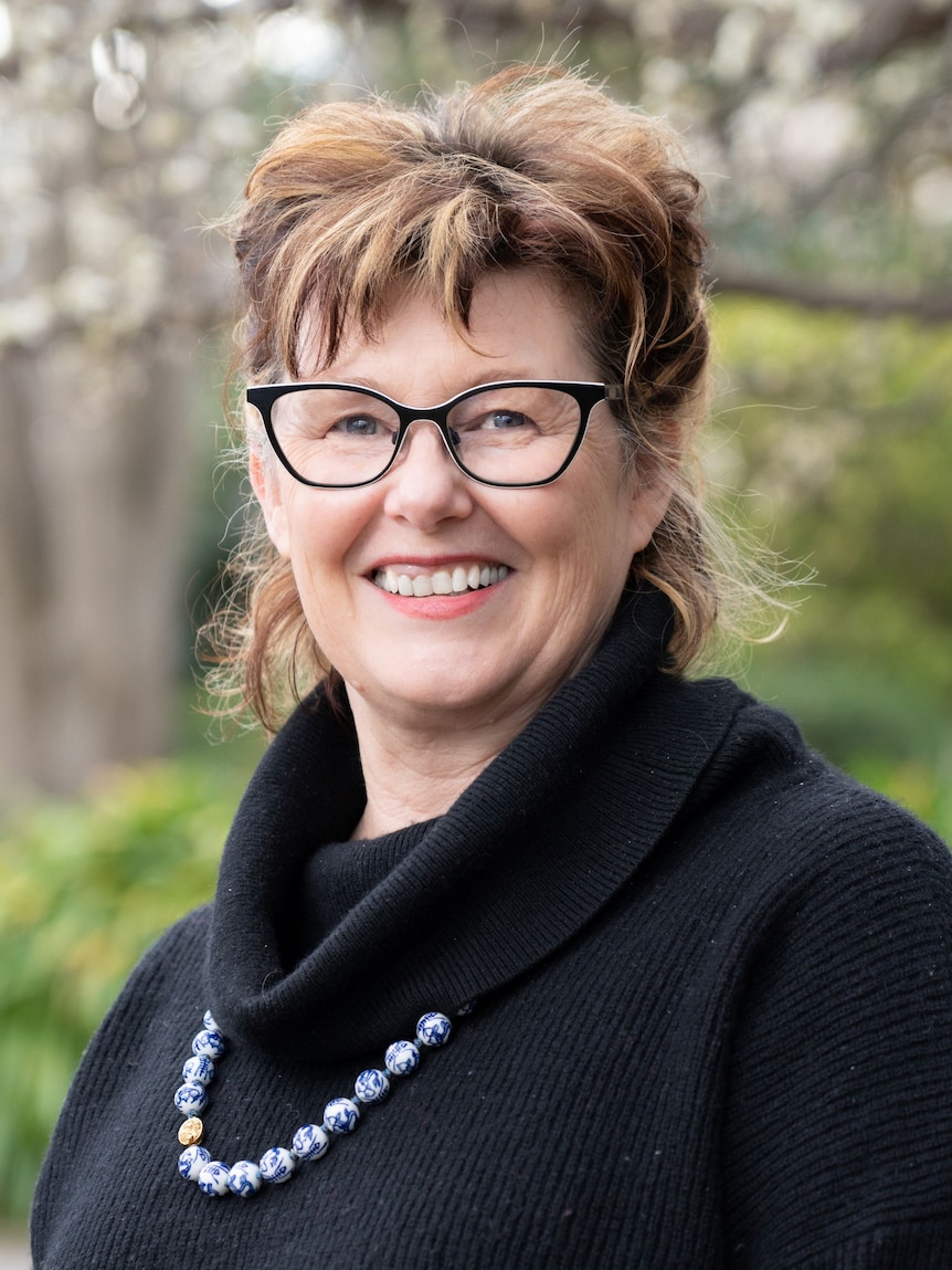 A woman with short brown hair and glasses, wearing a black cowl-neck top and a necklace