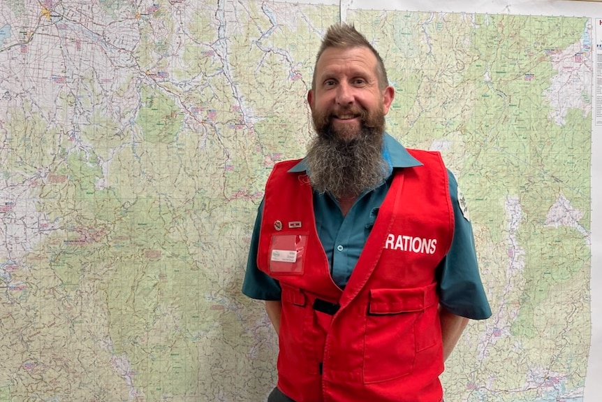 A man with brown hair and a long beard wearing an operations controller vest stands in front of a map.