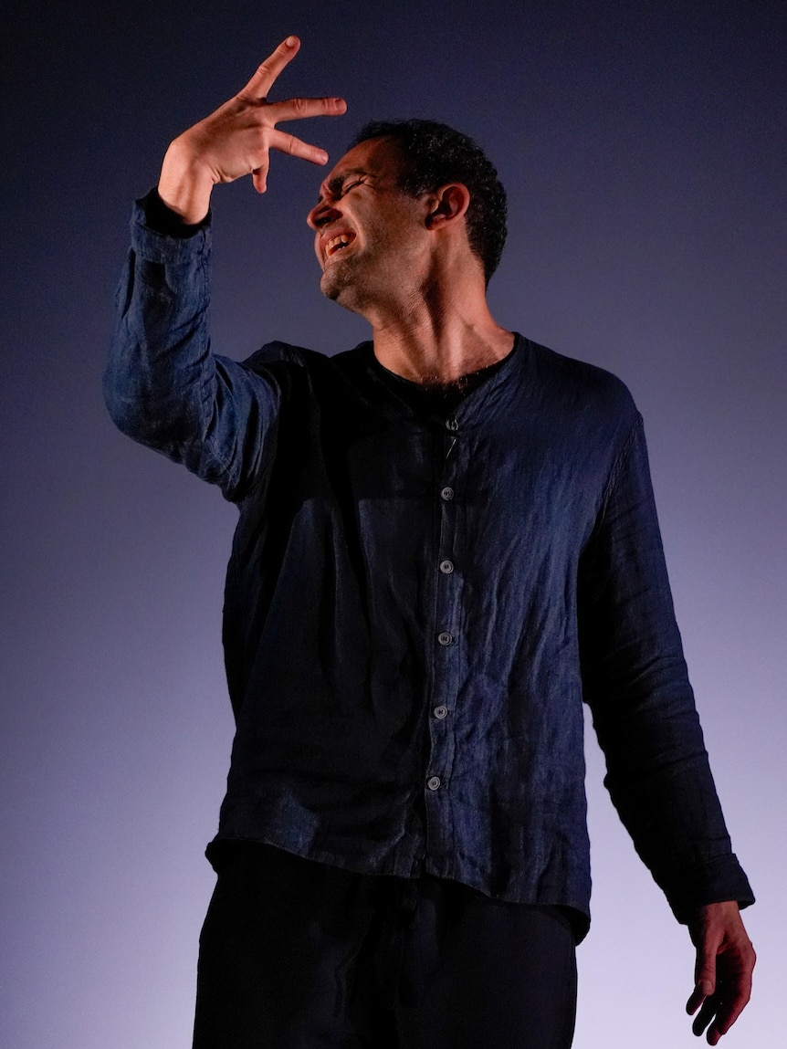 Khalid Abdalla, an Egyptian British man, 45, in blue linen, has an anguished expression, one arm raised, fingers splayed.