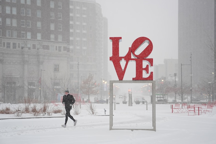A person jogging while it snows next to a red sign spelling out 'LOVE'.