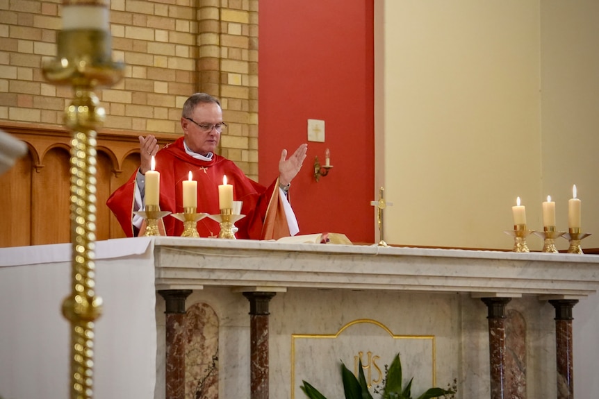 A clergman in robes gestures as he speaks from an altar with candles upon it.
