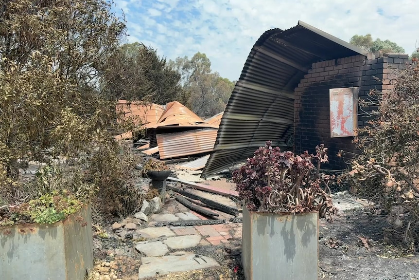 The crumpled tin roof of a house lies on the ground next to blackened brick walls.