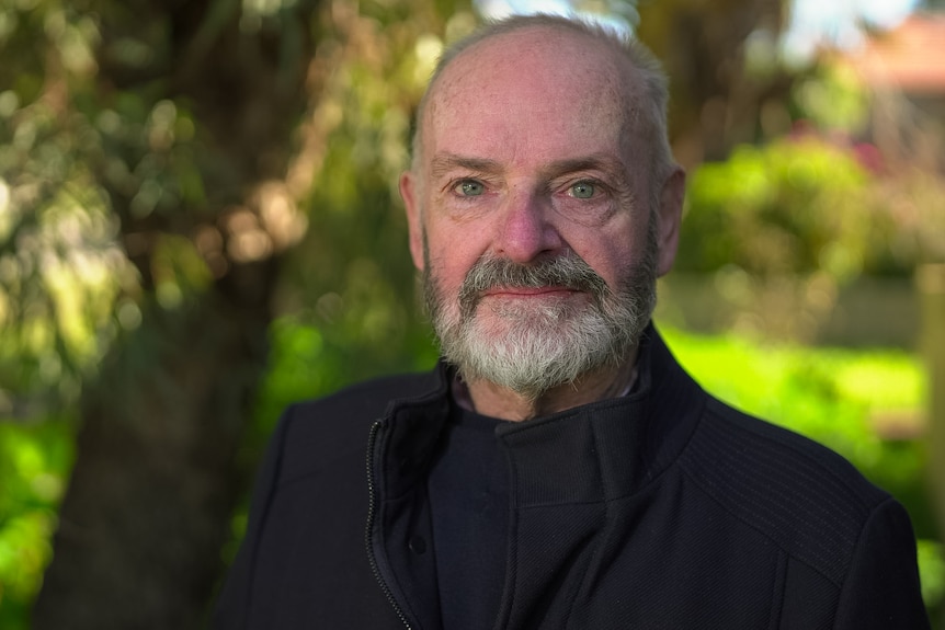 A bald man wearing a coat smiles at the camera in front of a lush backyard.