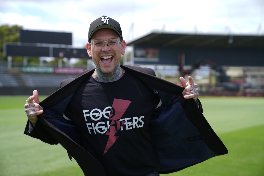 A man in a band tshirt and blazer smiles and poses with his tongue stuck out.