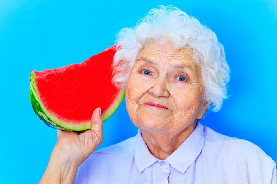 Elderly person smiling while holding a slice of watermelon up to their ear playfully, set against a plain background