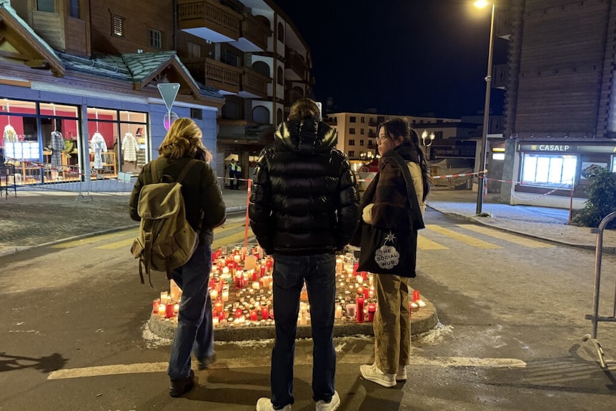 Three people stand in front of a memorial with candles and flowers set up on a street.