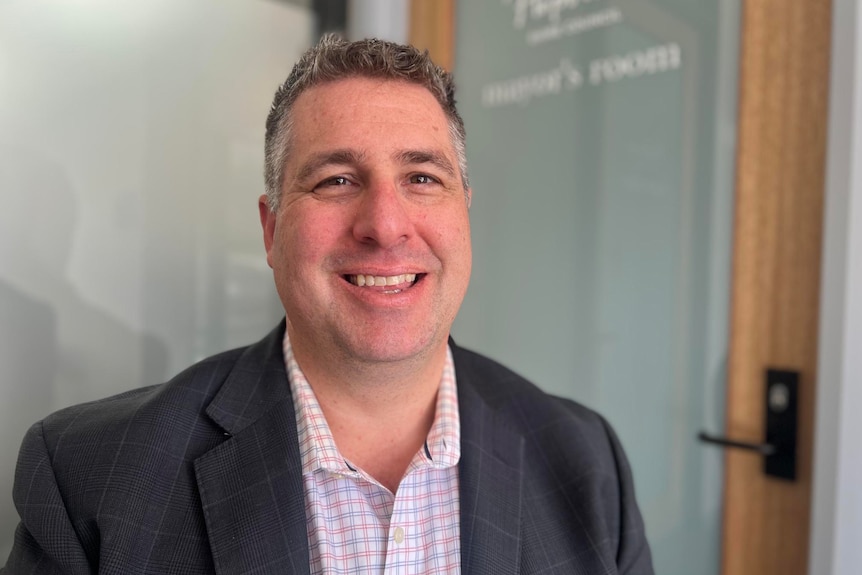 Grey haired white man in checkered shirt and dark blazer, sitting in front of door that says Mayor's room. 