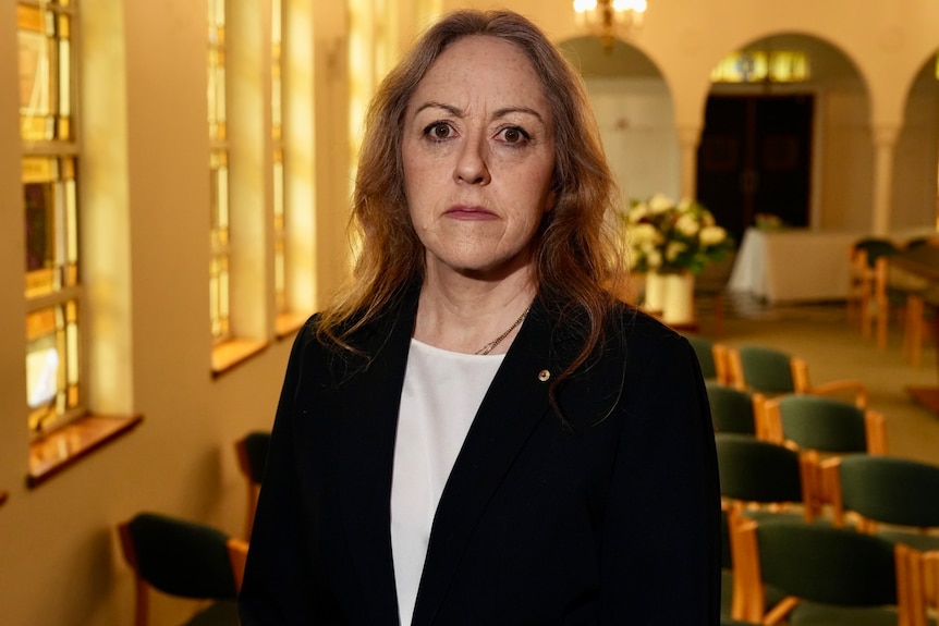 Rabbi Jacqueline Ninio standing inside a synagogue in a navy suit.