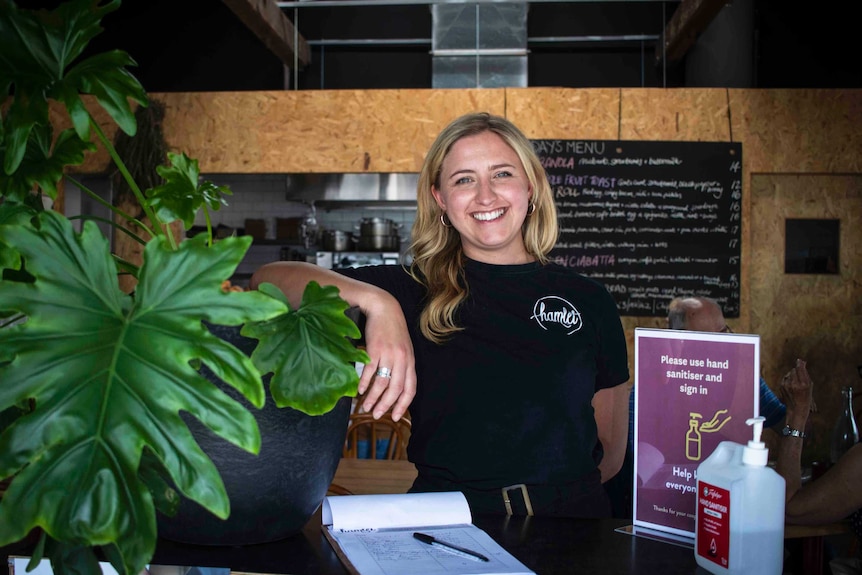A woman stands in a cafe smiling, her arm rests on a pot plant.