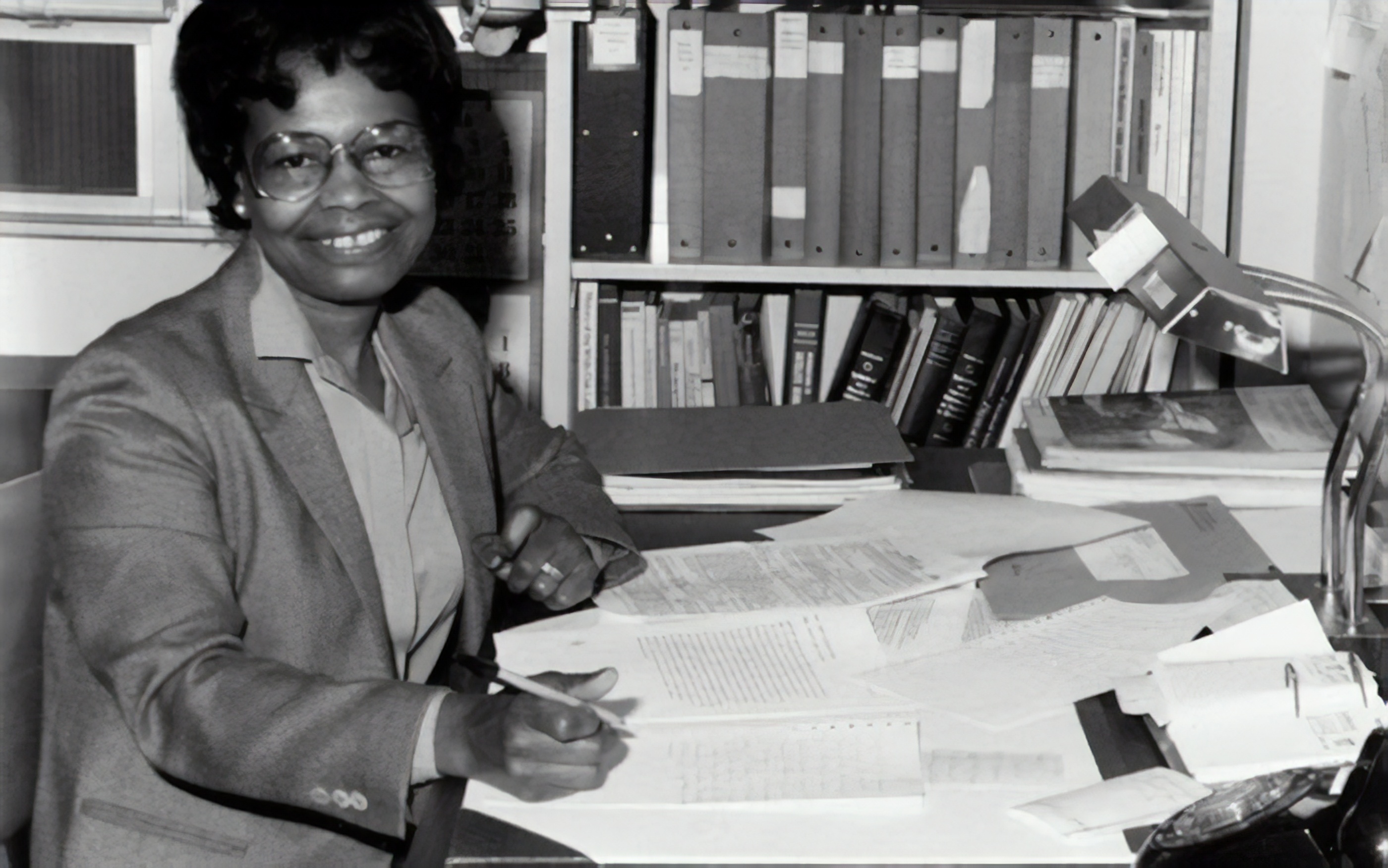 A woman in glasses and a blazer sits at a desk covered with papers, writing, with shelves of books and binders in the background.