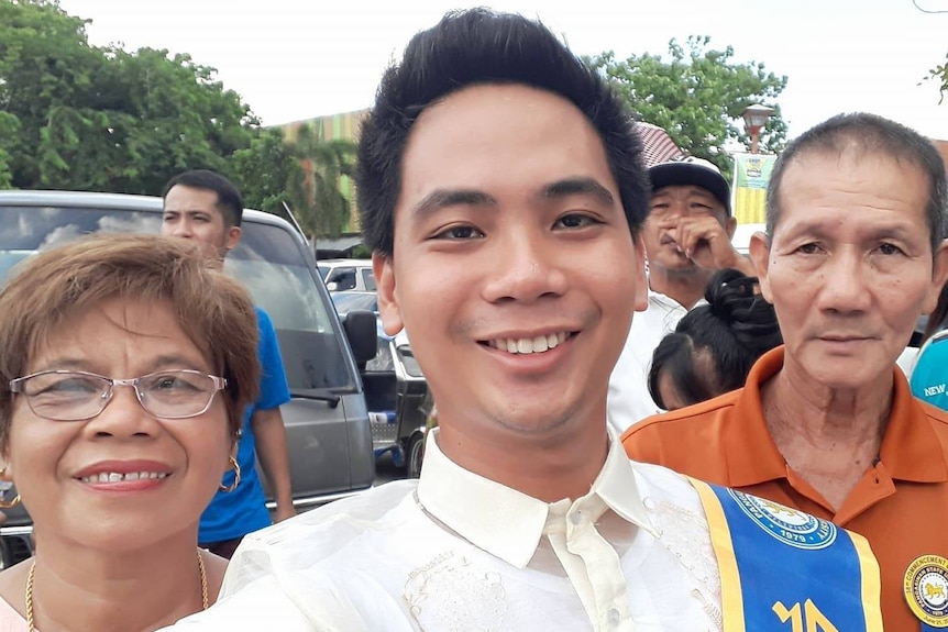 A young man stands between his mother and father. All three are smiling.