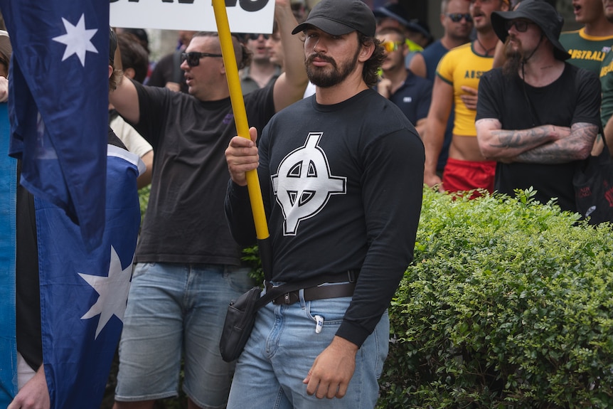 A man in a black shirt with a white Celtic cross, holding a sign.