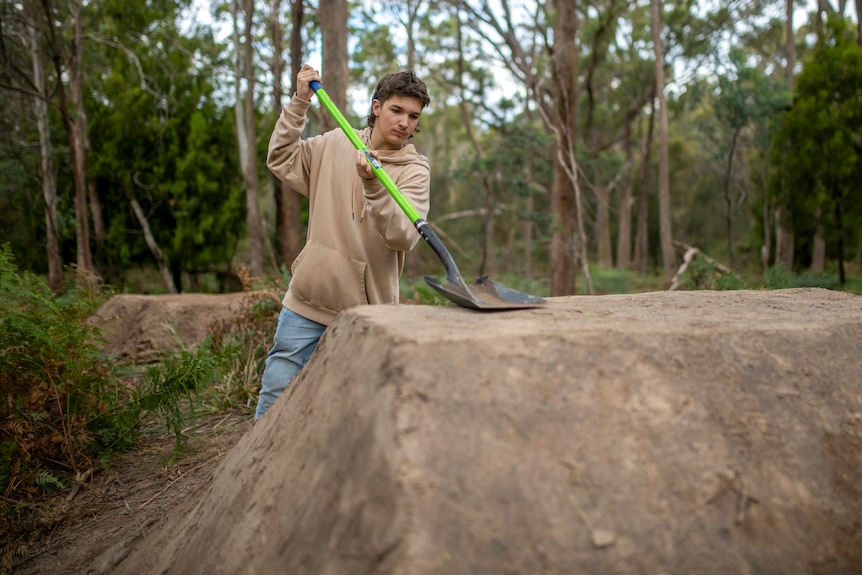 A teenager in a beige hoody and jeans pats down dirt bike jumps with the back o a large shovel in the bush.