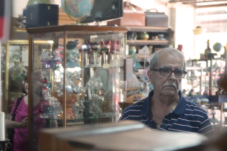 A older man stands in an antique shop surrounded by antiques.