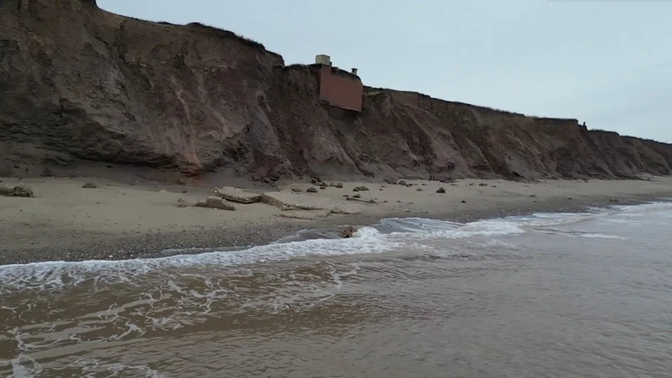 Waves are seen in the foreground and beyond that the tall brown cliffs where the brown brick bunker is perched.