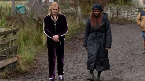 Sony Pictures Jack O'Connell and Nia DaCosta on the set of 28 Years Later: The Bone Temple - he wears a purple velour tracksuit and gold chains and has shoulder length blond hair, she has a long black padded coat and short boots - they are looking down and walking outside on a muddy path 