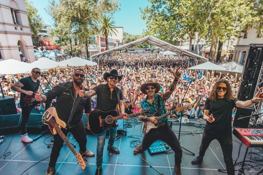 A band stand on a stage posing for the camera, behind them a crowd cheers