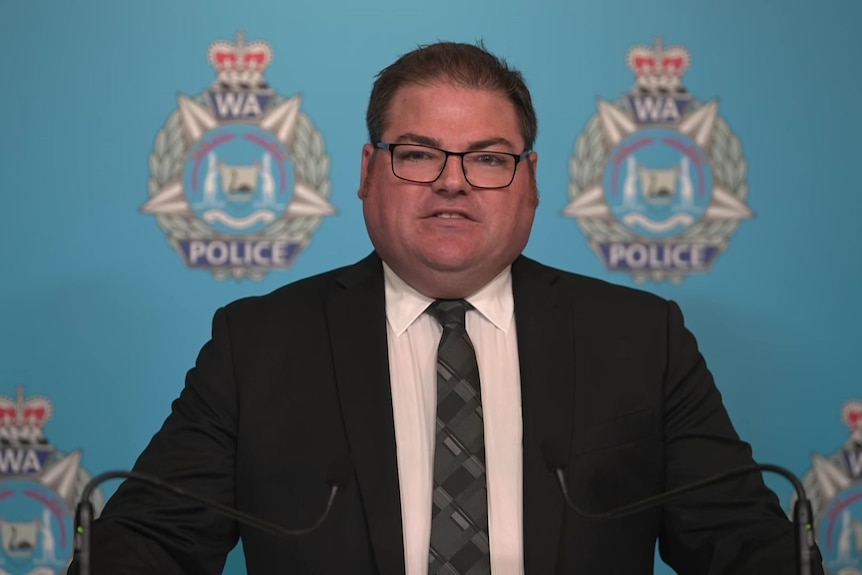A police officer wearing a suit stands in front of a WA Police backdrop.