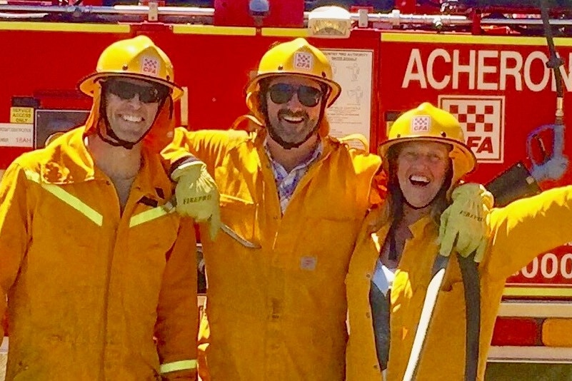 Three people smiling in yellow fire fighting gear, including helmets, stand in front of a fire truck.