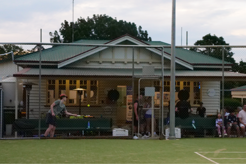a wide shot of the clubhouse with people sitting out the front and a teenage boy bouncing a ball on his racquet