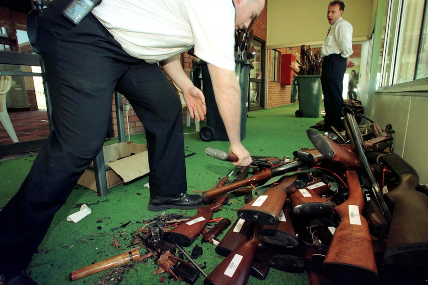 A man stoops to lay a shotgun on a pile of other guns.
