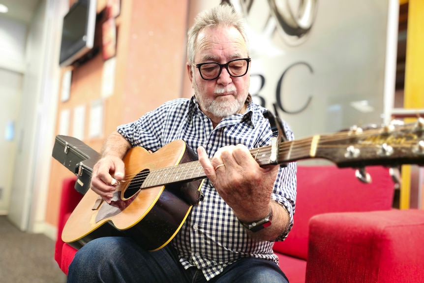 Older man wearing a blue checkered shirt plays the guitar while sitting on a red couch.