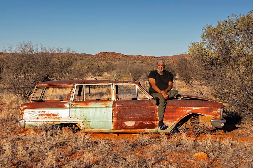 An Aboriginal man sits on the bonnet of rusted car abandoned in the desert.