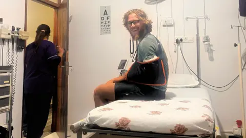 Olly Hargreaves Olly sits on a hospital bed, his left arm is in a sling, he has collar-length blonde hair and a long ginger beard. He smiles at the camera