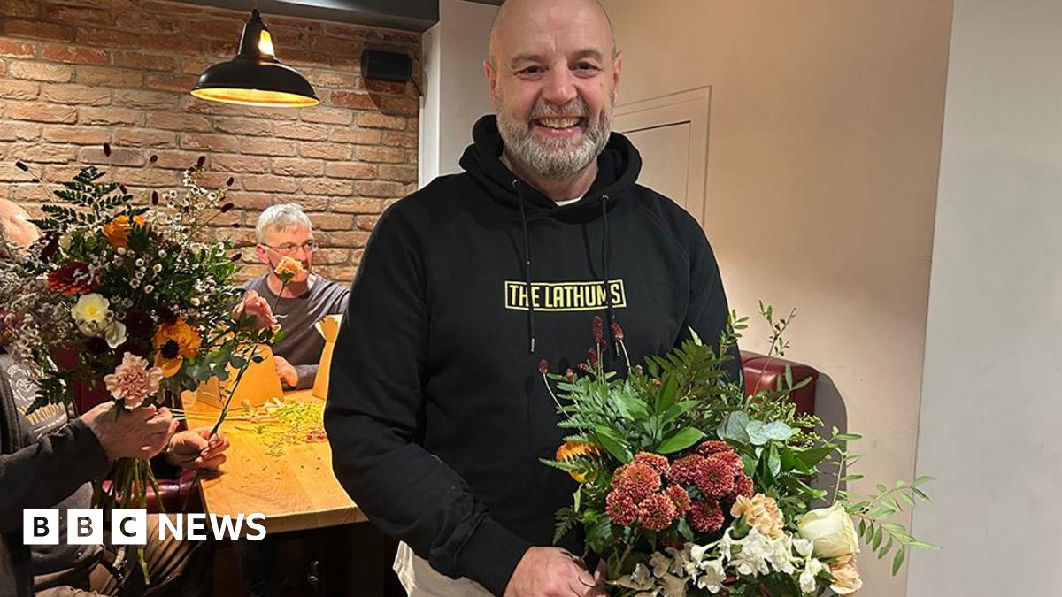 Stuart Leatherbarrow, who is bald and has a short grey beard, stands in front of a table where men are arranging flowers. He is wearing a black hoodie and holding a large beautiful bouquet.