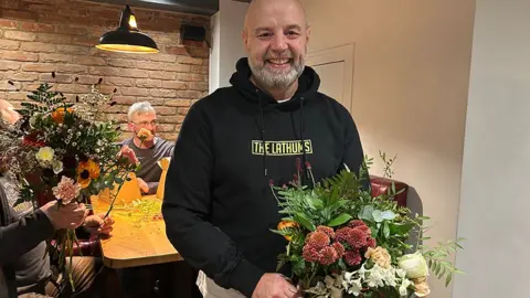BBC Stuart Leatherbarrow, who is bald and has a short grey beard, stands in front of a table where men are arranging flowers. He is wearing a black hoodie and holding a large beautiful bouquet.