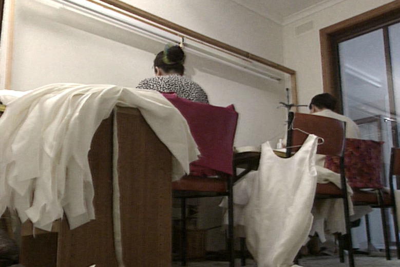 Two women seen from behind with white dresses draped on a bench next to them. 