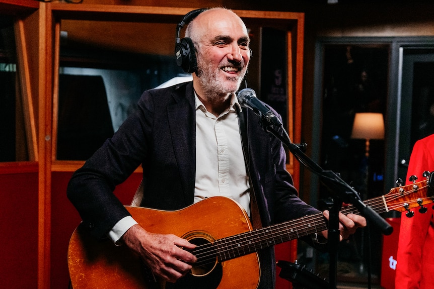 A man with headphones on holds a guitar in a studio. He is smiling.
