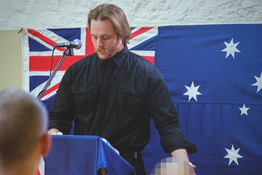 Craig Johnson stands at a lectern draped in an Australian flag, in front of an Australian flag hanging on the wall