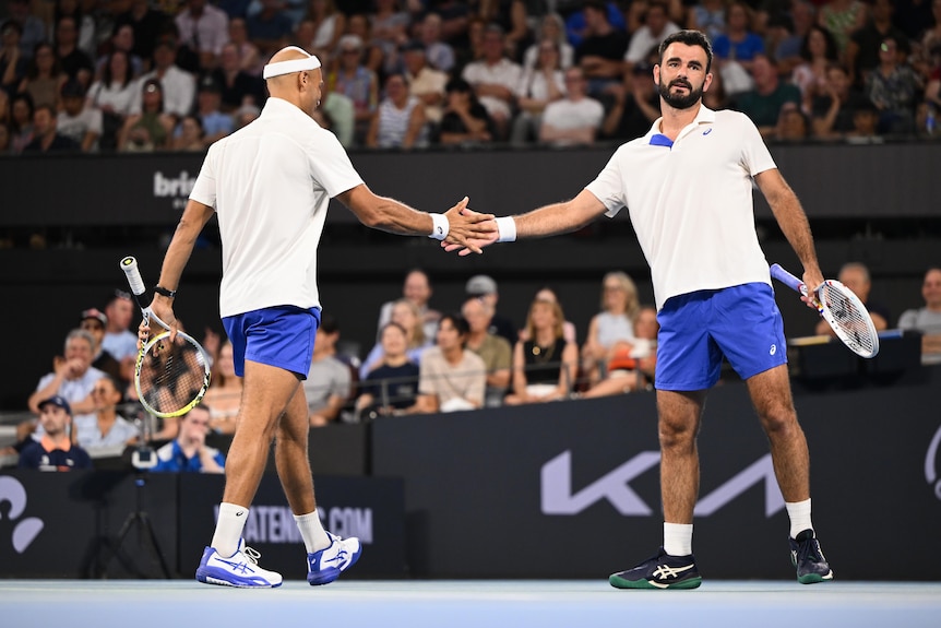 Two male tennis players wearing white and blue high five on the court