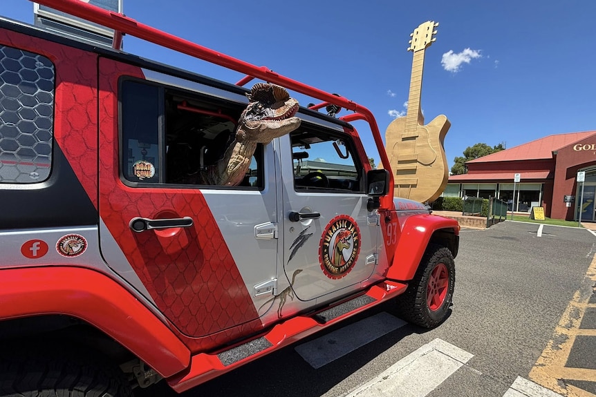 A model dinosaur pokes its head out the back window of a Jurassic Park vehicle in front of a giant guitar sculpture