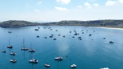 BBC Image shows yachts on calm blue waters with golden sand and green coastline of the island of Alderney.