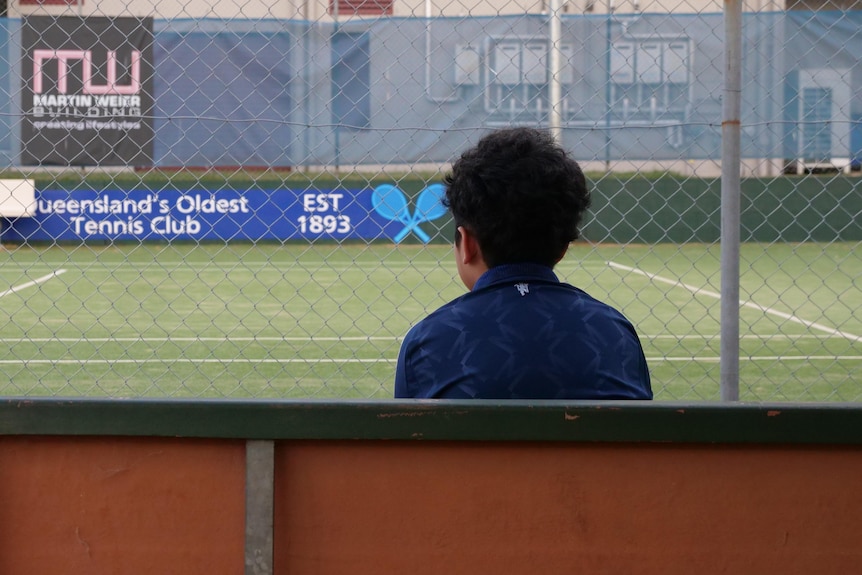 the back of a young boy sitting on a bench looking at a tennis court